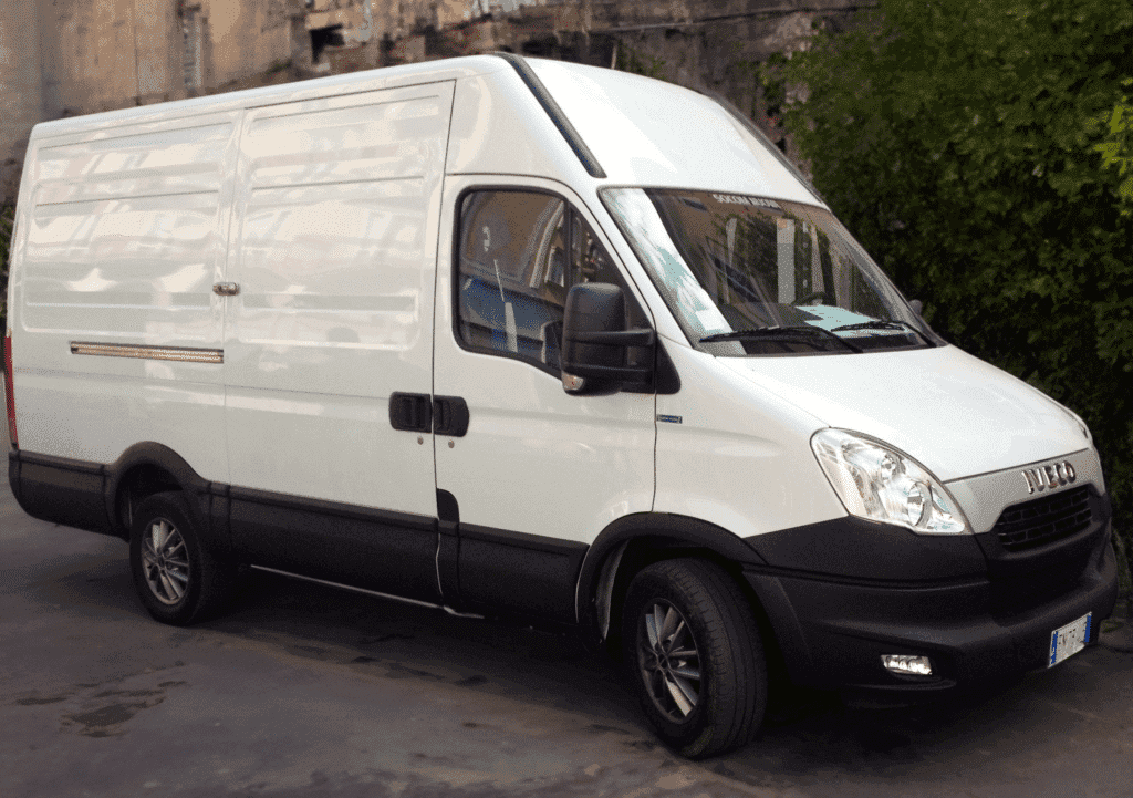 A white delivery van parked on a concrete surface, featuring a sliding door and pronounced black wheels, with shadow reflections on its side.