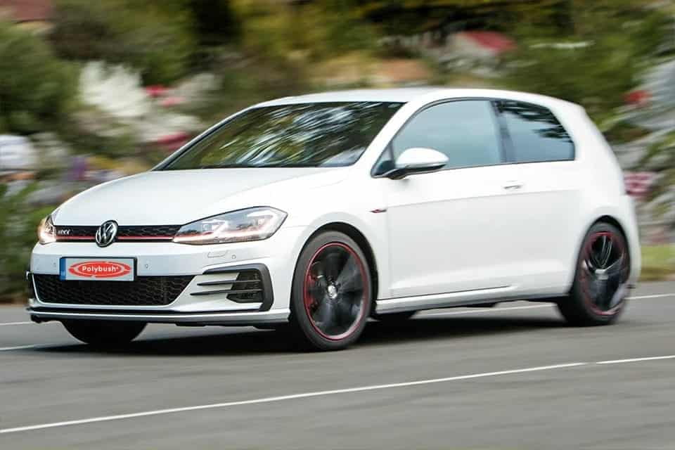 A white Volkswagen Golf GTI moving swiftly on a blurred road, exhibiting its sporty design and dynamic performance. The car features distinctive red trim on wheels and a black roof.